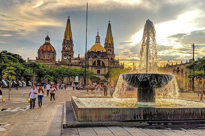 Fountain sprays water in front of Guadalajara Cathedral, Mexico, with people walking on a plaza at sunset.