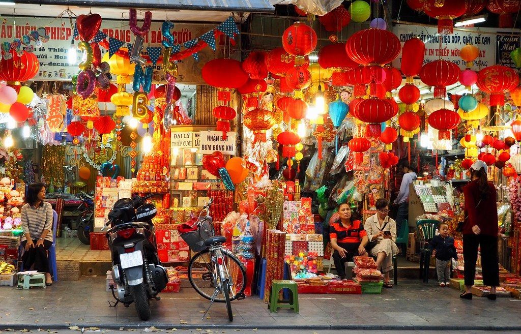 A street vendor stall in Vietnam overflowing with red lanterns and decorations. People shop and browse in the festive, brightly lit space.