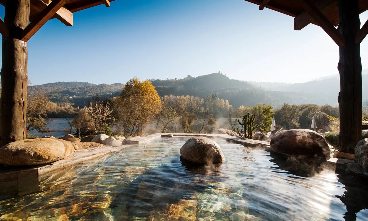 Outdoor hot spring pool with mountain view, under wooden shelter; water, rocks, trees. Spain hot springs