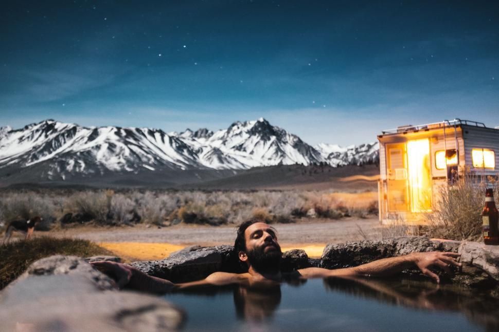 Man in hot spring, arms outstretched, under starry sky with snowy mountains and lit trailer. hot springs Europe