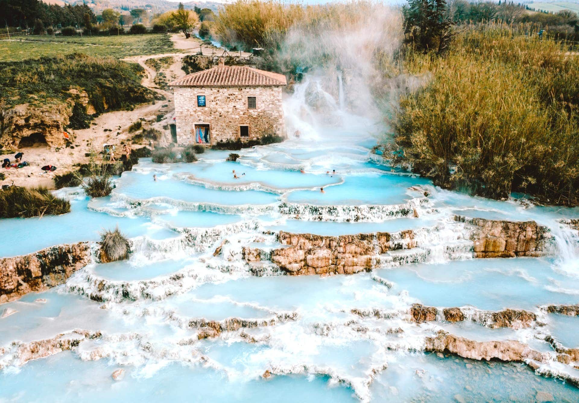 Cascading turquoise thermal pools with a stone building, surrounded by greenery and steam.