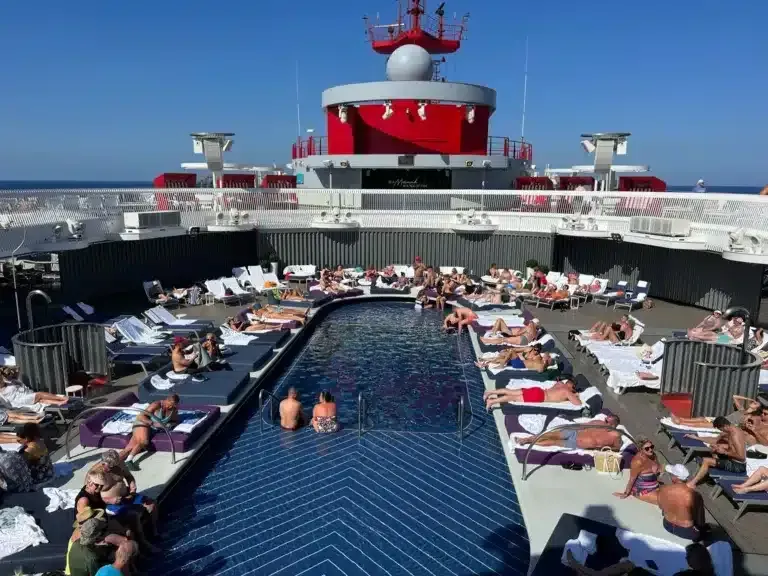 People relax on lounge chairs around a dark blue chevron-patterned swimming pool on a cruise ship deck.