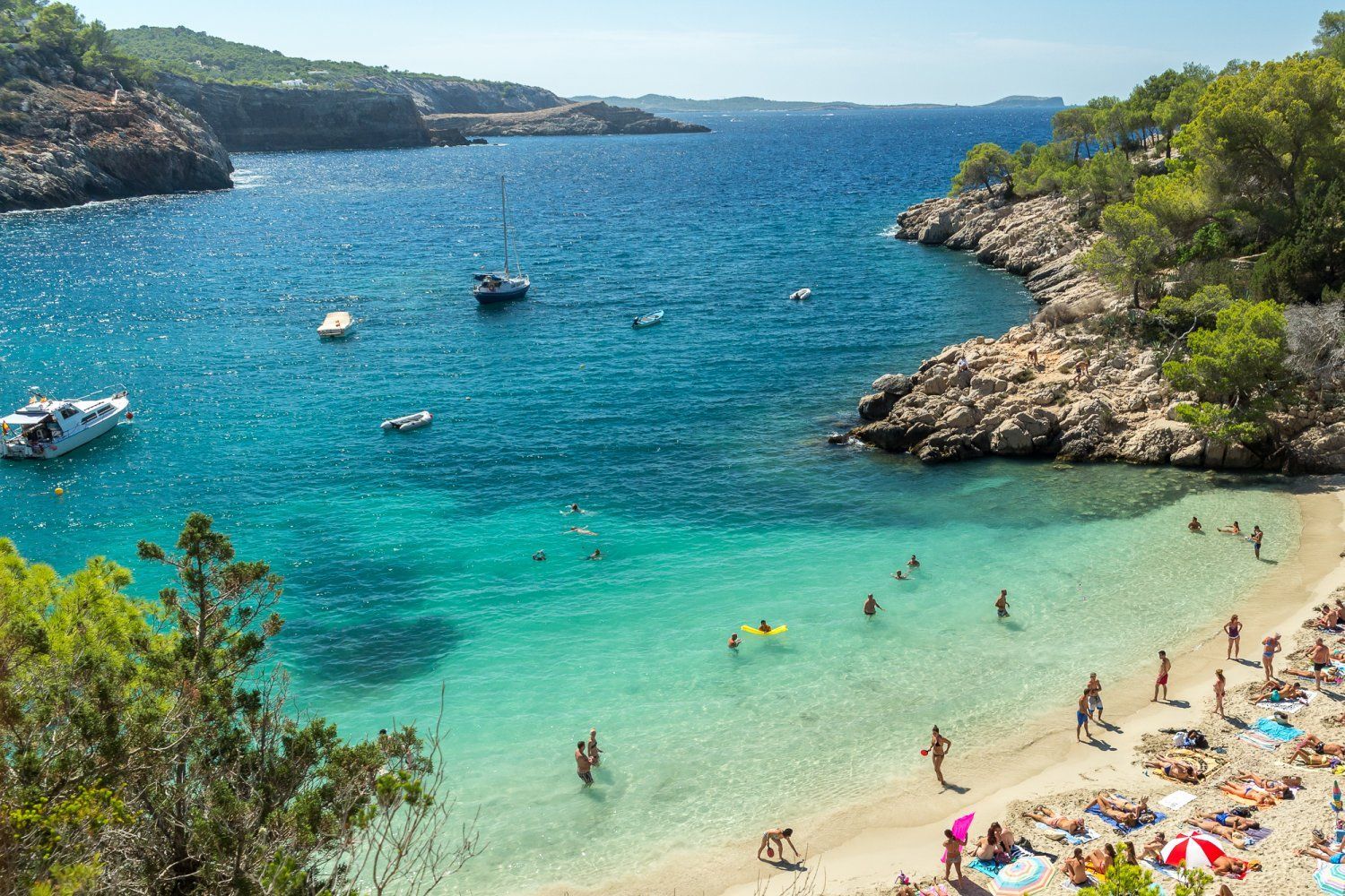 Scenic beach cove with turquoise water, boats, and people swimming and sunbathing.