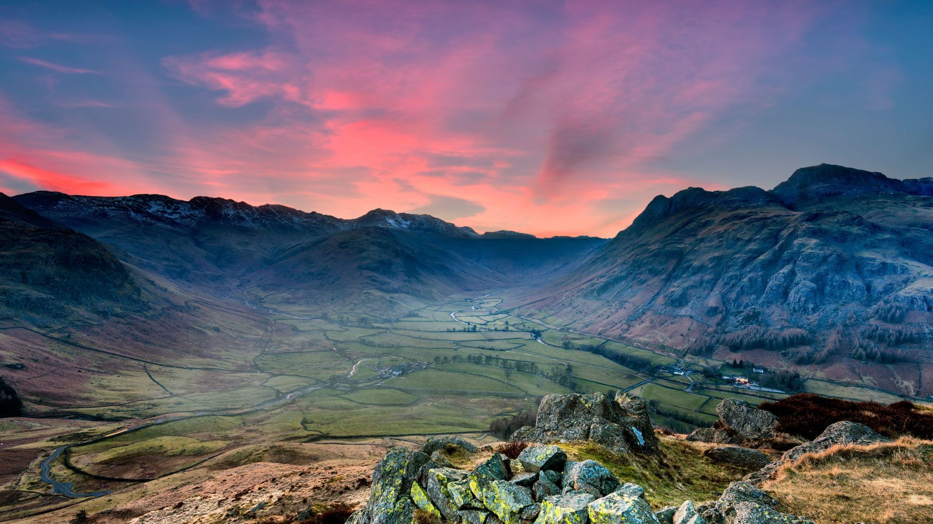 Valley view at dusk, mountains silhouetted against a fiery pink and orange sky.