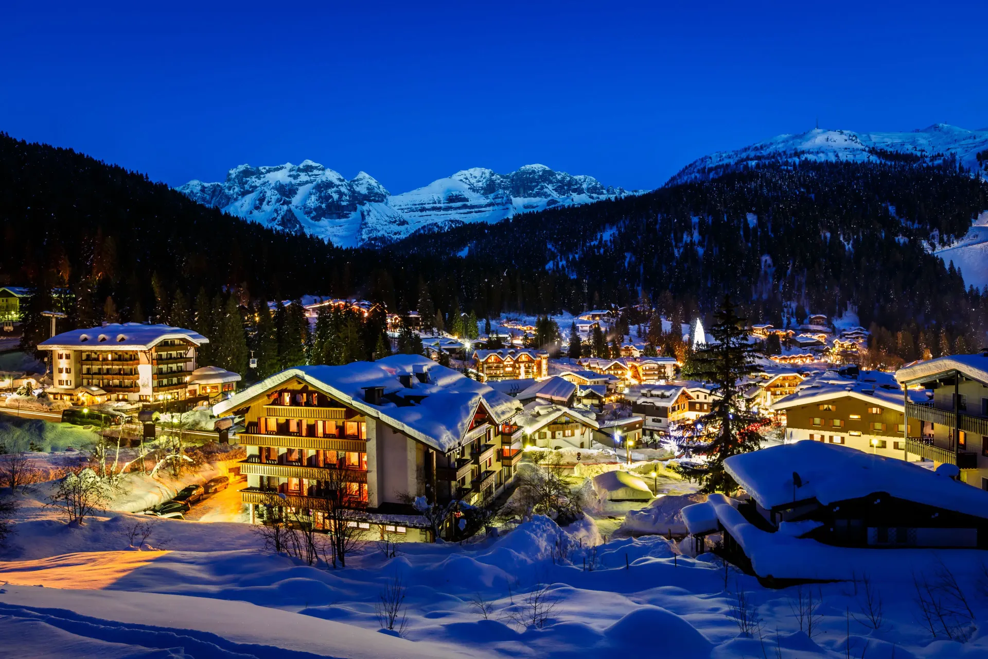 Snow-covered village nestled in a valley with lit buildings and snowy mountains under a dark blue sky. Madonna di Campiglio, Italy
