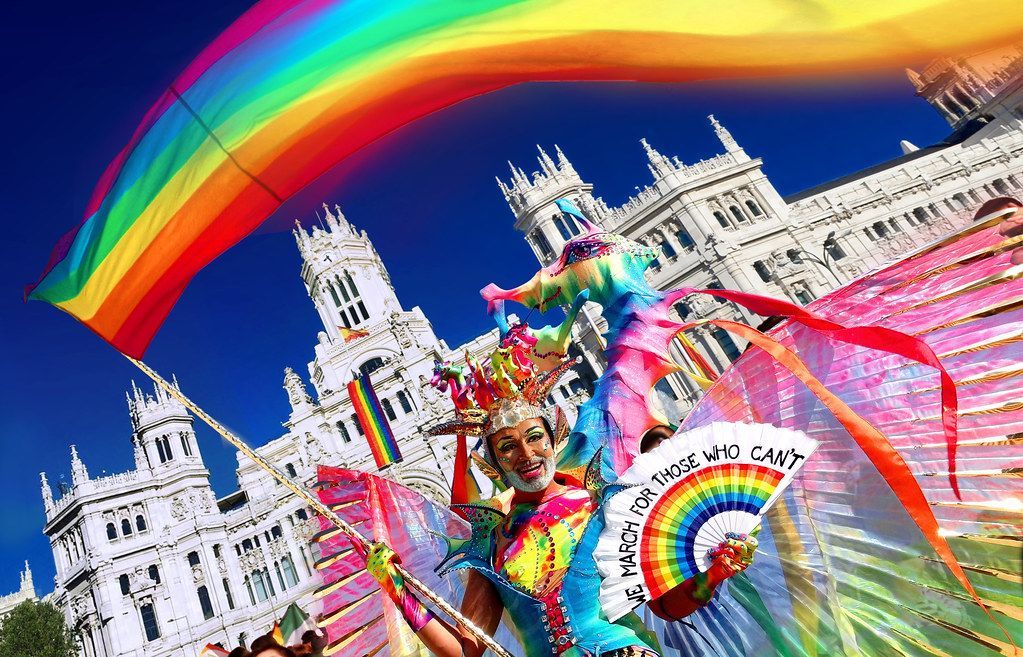 Person in colorful costume with rainbow fan at Madrid Pride, rainbow flag and ornate building backdrop.