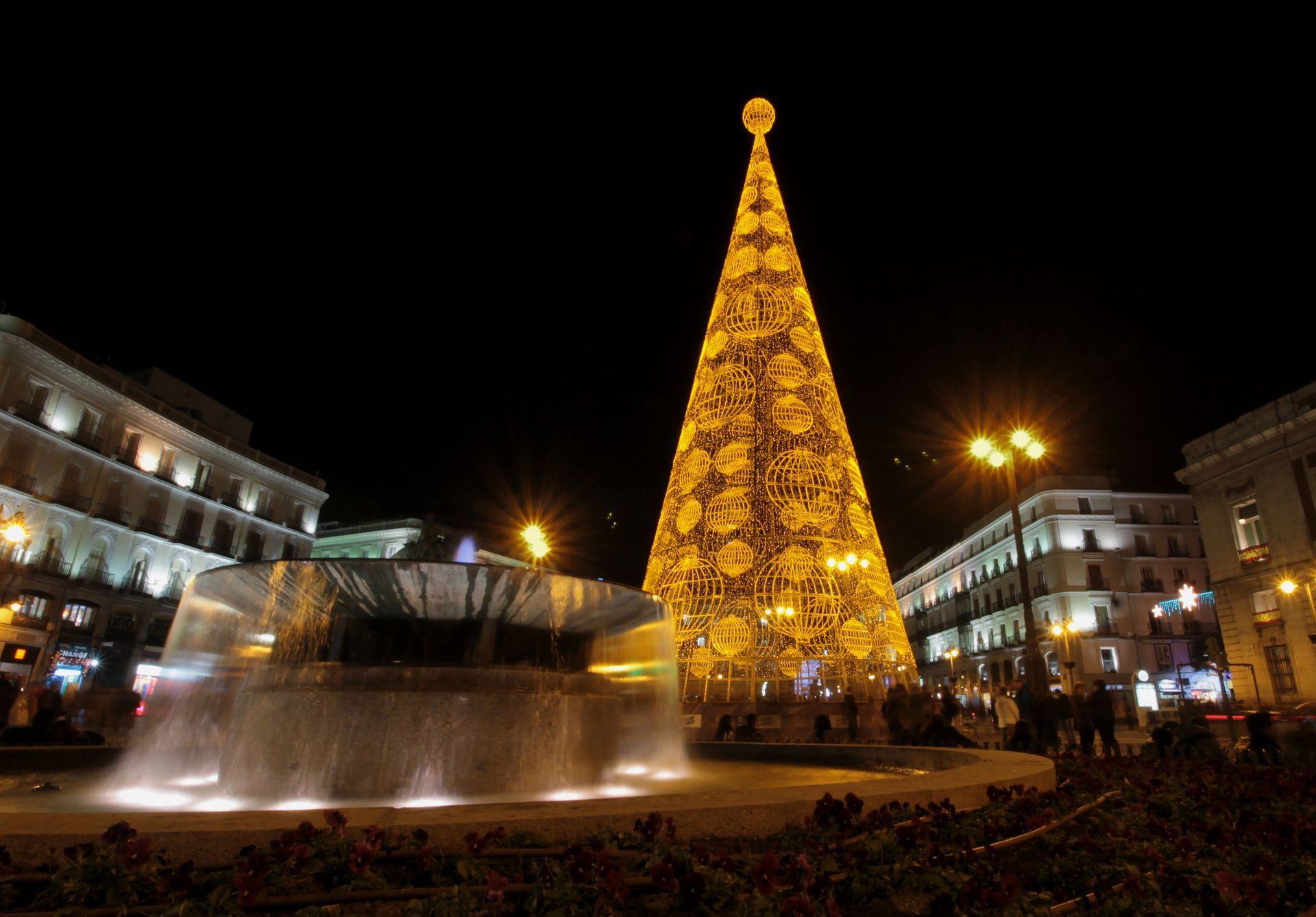 Night scene of a lit Christmas tree in a plaza with a fountain. Buildings and lights frame the view. Madrid