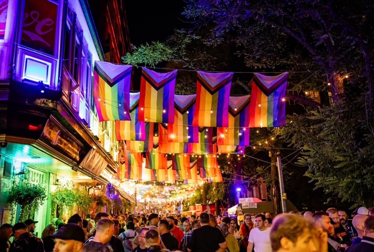 Nighttime street festival, rainbow and progress pride flags overhead, bustling crowd, colorful lights.
