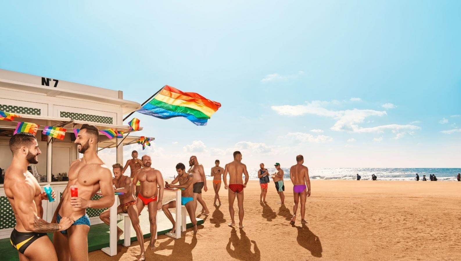 Men in swimsuits on a beach, gathered near a beach bar with a rainbow flag on a sunny day.