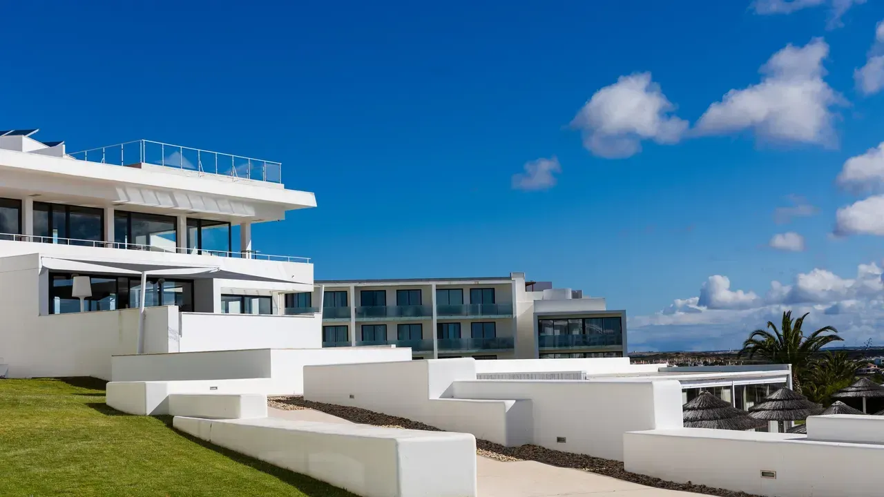 White modern buildings with blue windows against a bright blue sky and grassy hill. Memmo Baleeira Hotel