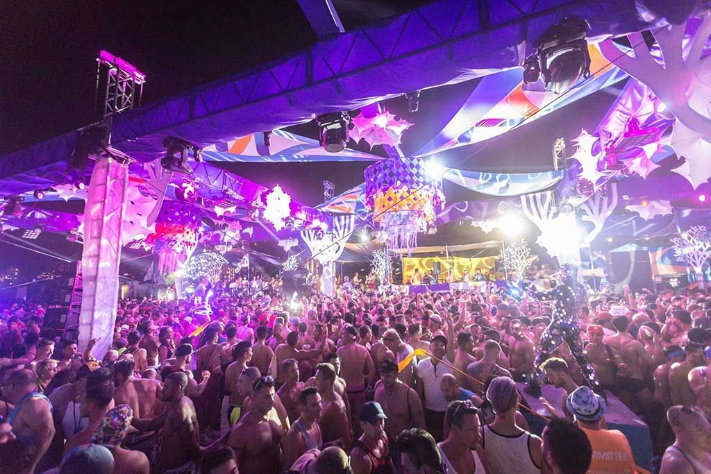 Miami Winter Party Festival (USA) A large crowd dances at an outdoor music festival under colorful lights and decorations. The scene is vibrant with purple and white hues.