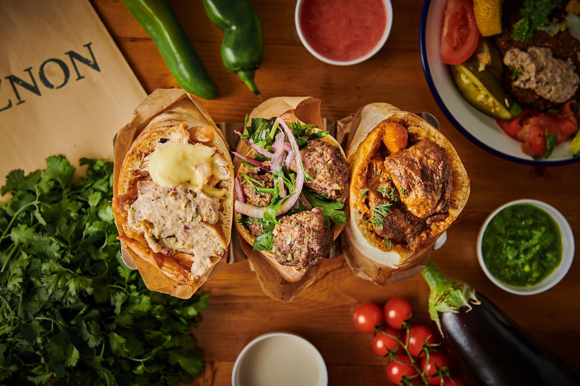 Three filled pita wraps on a wooden table, surrounded by ingredients and sauces. Miznon (near Le Marais)