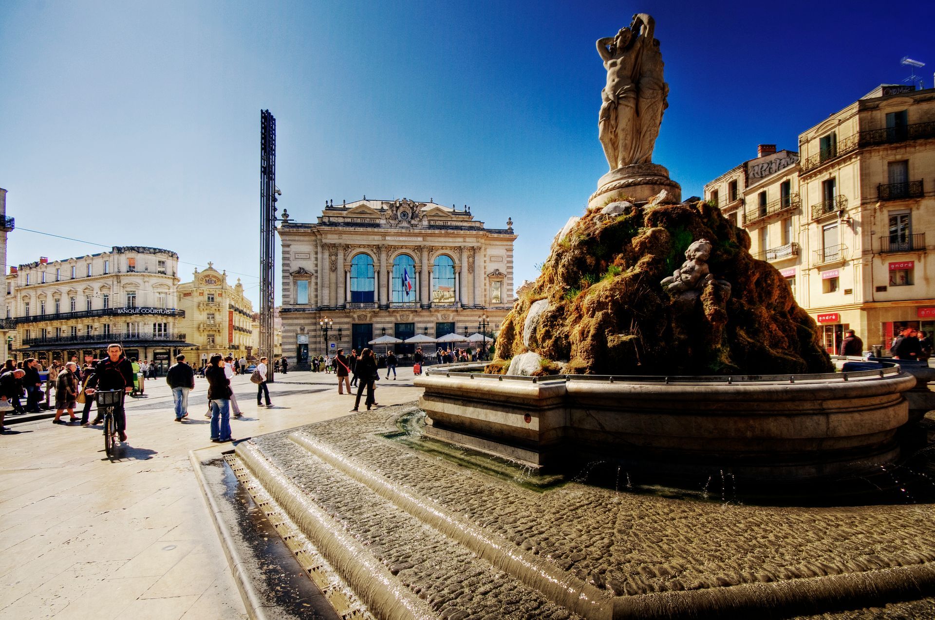 A sunny city square with a fountain and a large building with arched windows. People walking. Montpellier – France’s Southern Capital of Pride