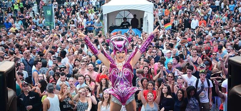 A drag performer in a sparkly pink costume with arms raised on a stage, cheered on by a large crowd. Montreal