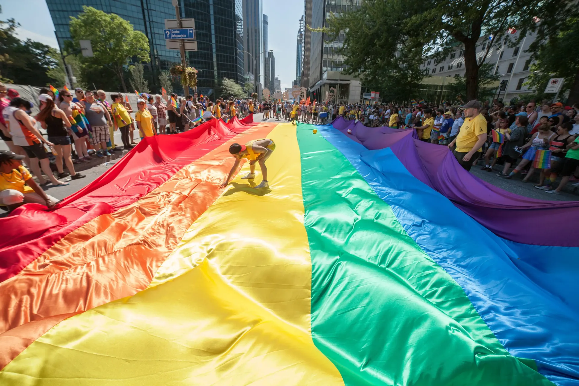 A giant rainbow flag carried down a city street during a Pride parade; people line the sides, cheering. Montreal Pride (Canada)