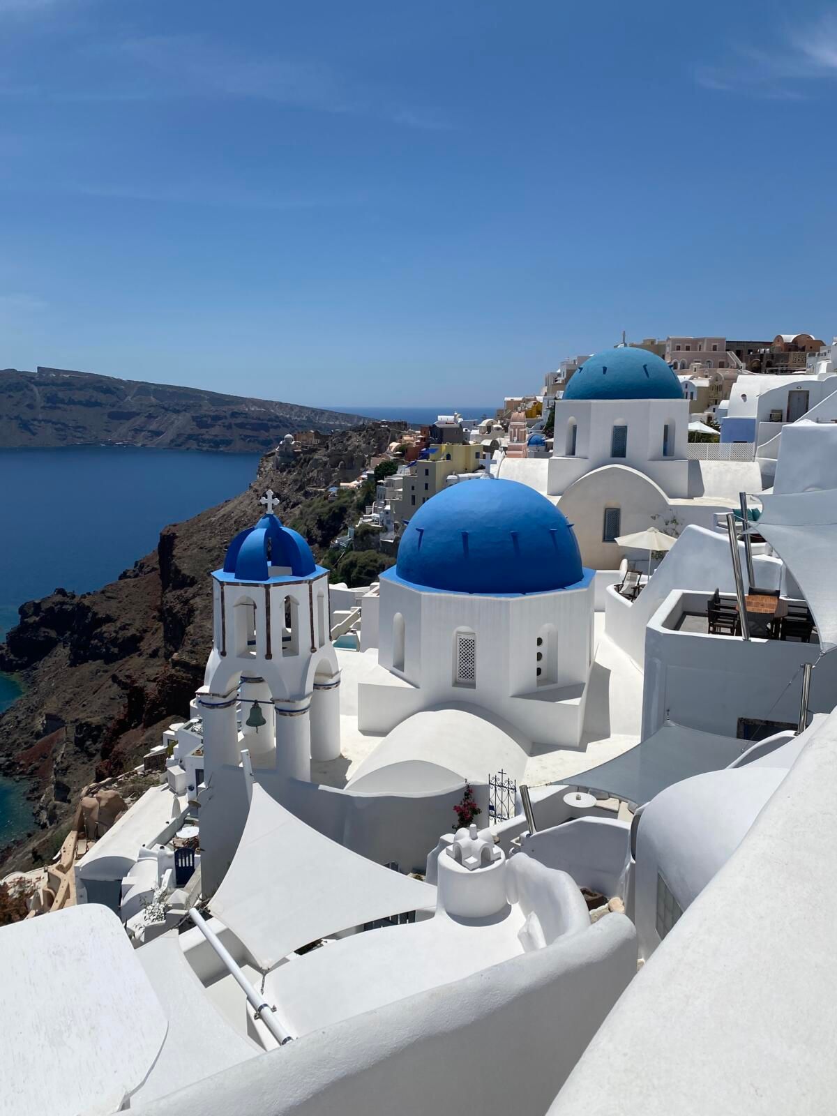 White buildings with blue domes on Mykonos cliffs, overlooking the Aegean Sea under a blue sky.