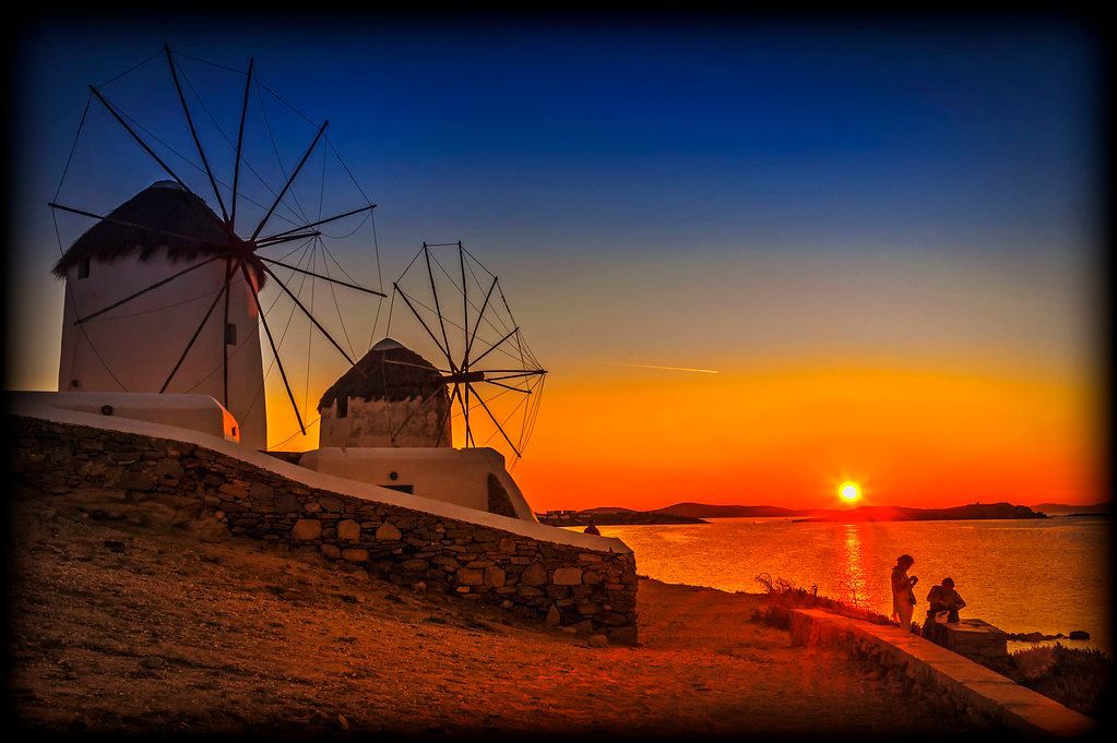 Windmills silhouetted against a vibrant orange sunset over the sea. People watch from the shore. Mykonos