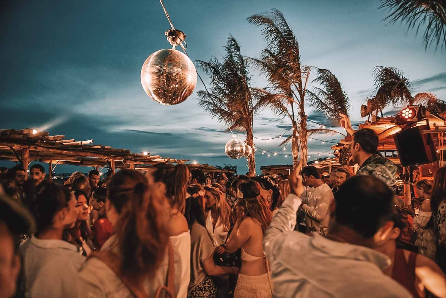 Party with disco balls, palm trees, and a crowd at dusk.