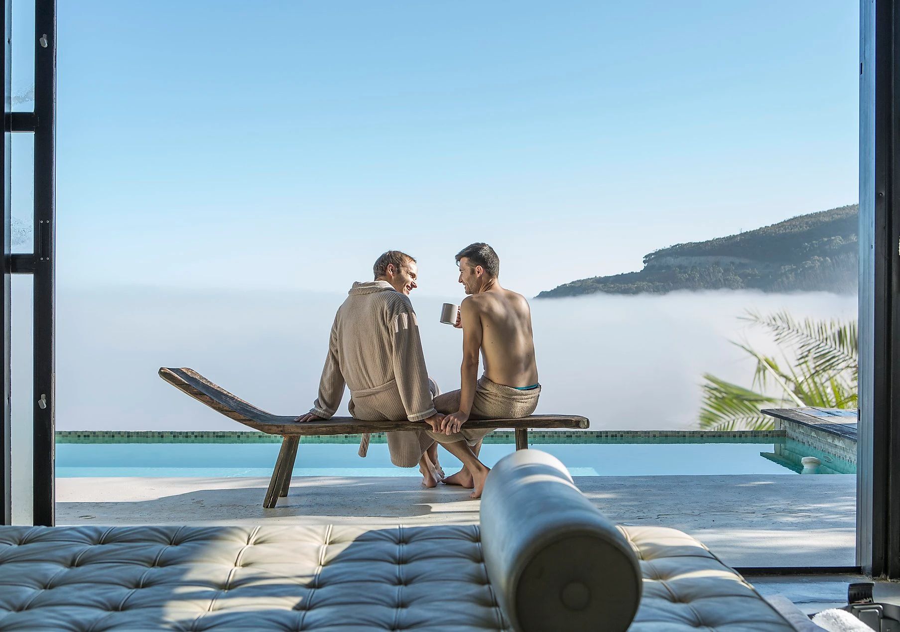 Two people relax on a chaise lounge near a pool, looking at a cloud-filled vista.
