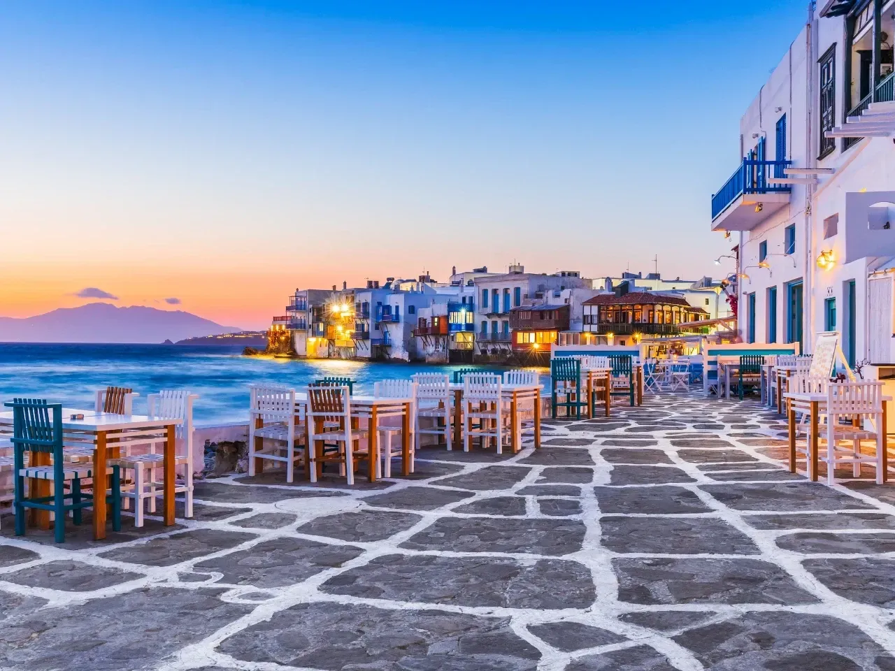 Restaurant tables and chairs by the sea in Mykonos, Greece, at dusk. White buildings and blue sky.