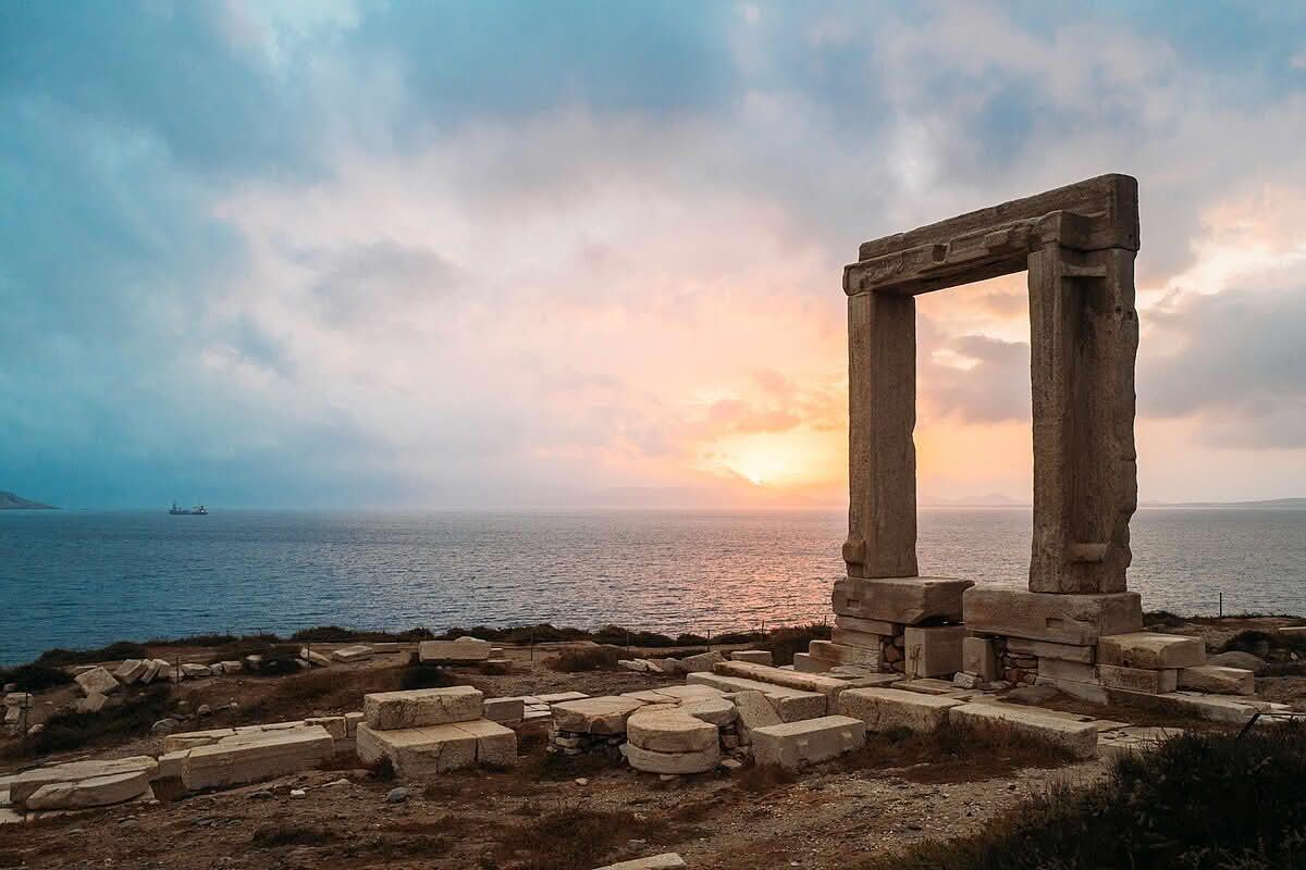 Ancient stone gate on a rocky cliff, overlooking the sea at sunset with a cloudy sky. Naxos