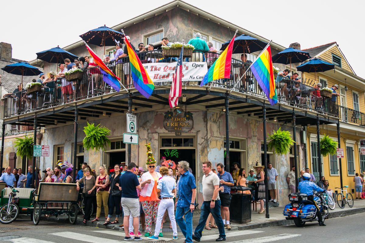 People gather outside a two-story building with rainbow flags, umbrellas, and balconies. New Orleans