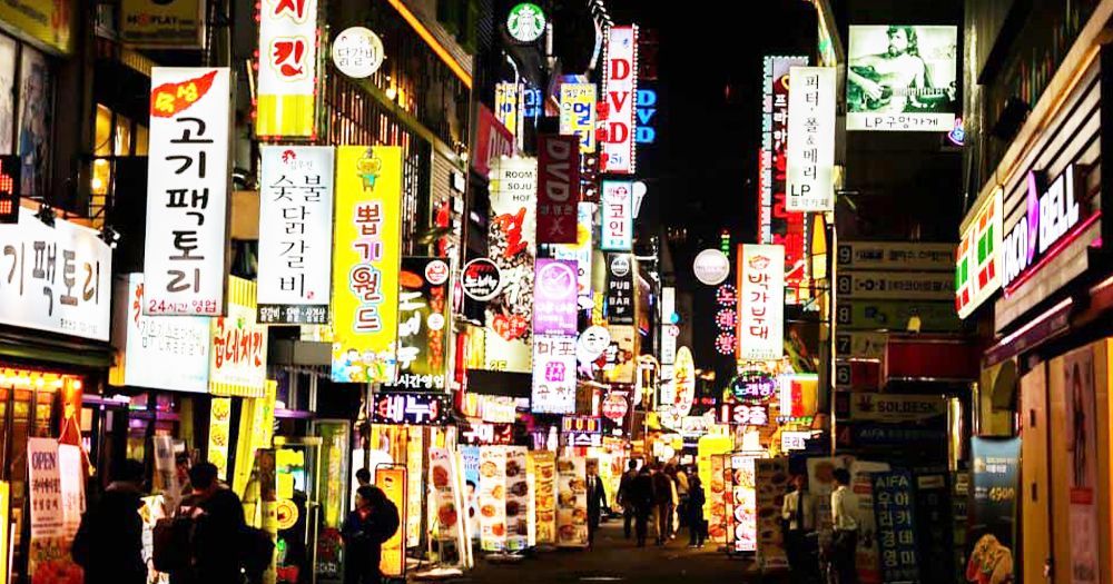 Nighttime street lined with brightly lit signs, bustling with people, likely in a city like Tokyo. Ni-chōme