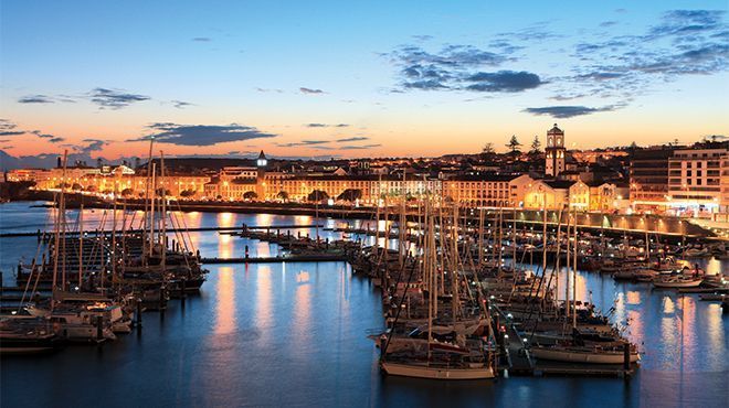 Boats docked in a marina at dusk, city lights in background reflect in the water. Ponta Delgada, Azores