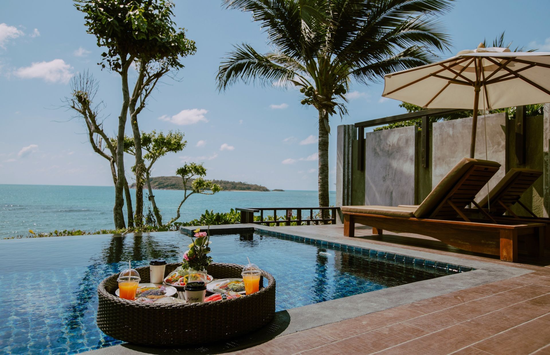 Infinity pool with a floating tray of food, ocean view, two lounge chairs, and palm trees.