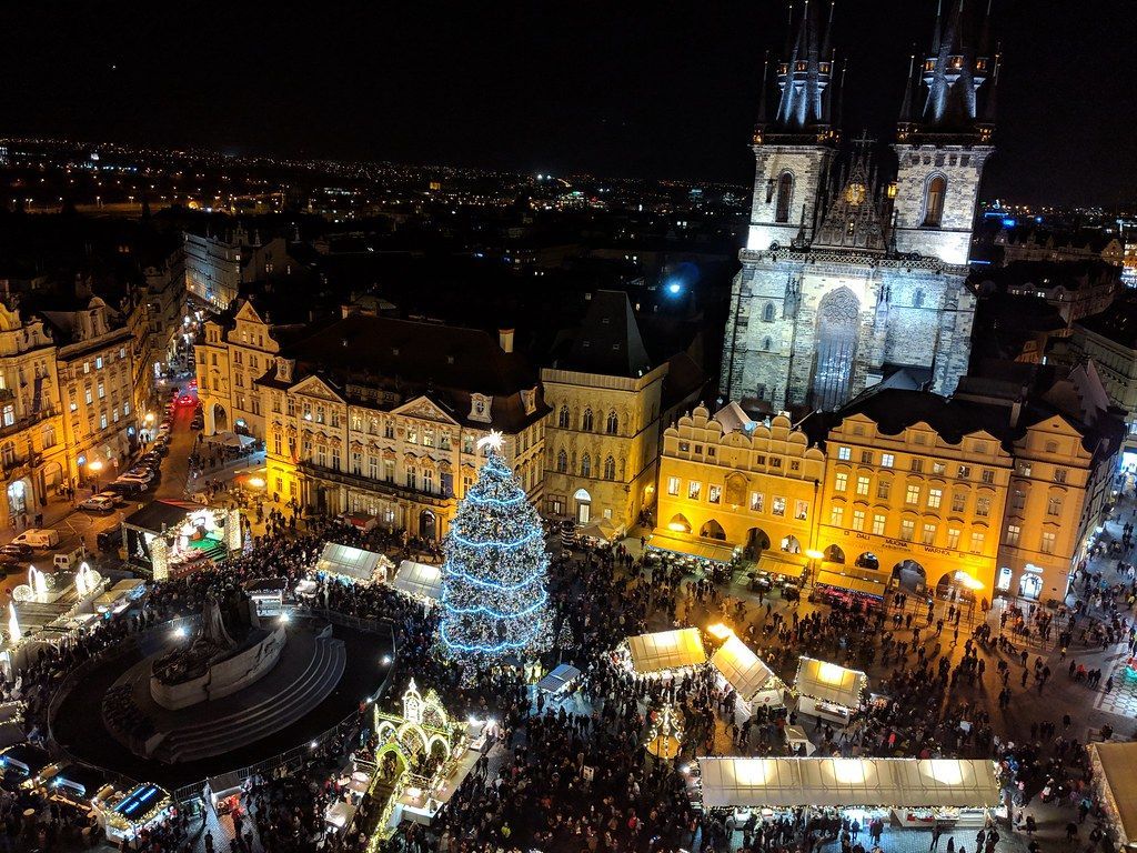 Nighttime view of Prague's Old Town Square, Christmas market, large illuminated tree, church towers, crowds, and buildings with lights.