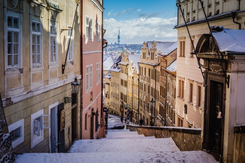 Snowy street in Prague, Czech Republic. Buildings line a sloped path; a tower is visible in the distance.