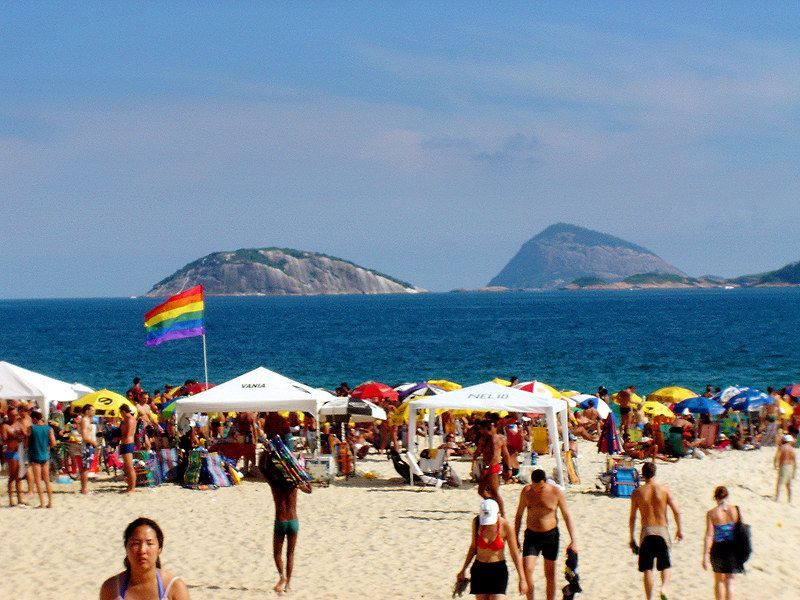 Beach scene with rainbow flag, crowded with people, ocean and islands in background. Praia 19, Costa da Caparica, Portugal