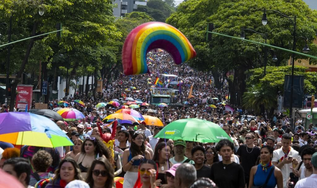 Crowd marching in a pride parade beneath a rainbow arch, with colorful umbrellas and trees lining the street