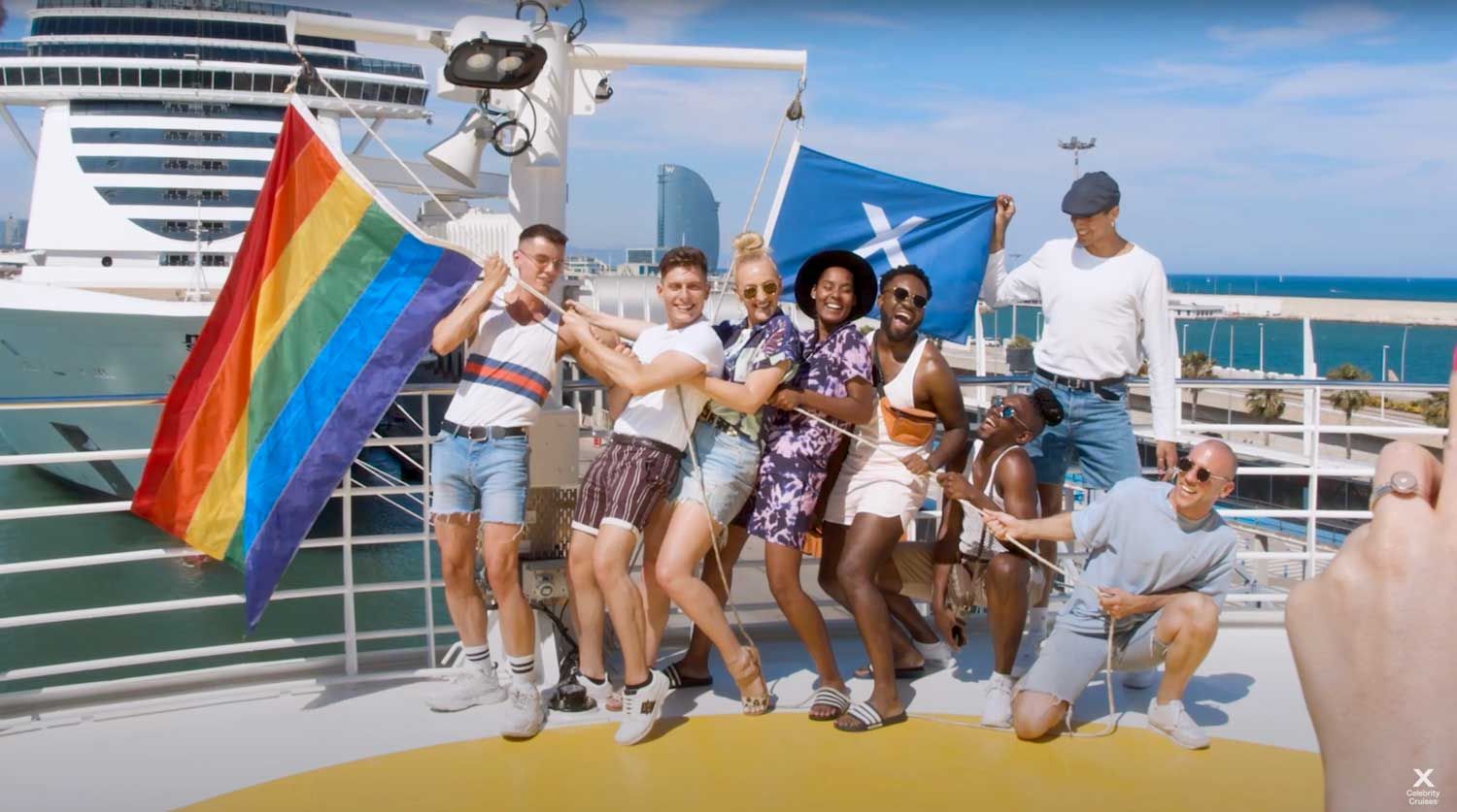 A group of people poses on a cruise ship deck holding pride and blue flags against a bright sky.