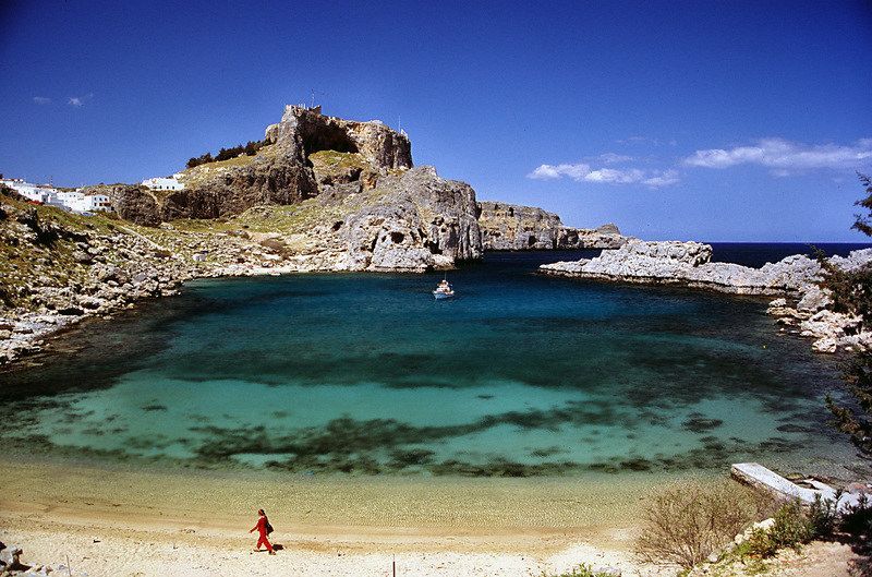 Bay with clear turquoise water and rocky cliffs under a bright blue sky. A person walks on the beach.