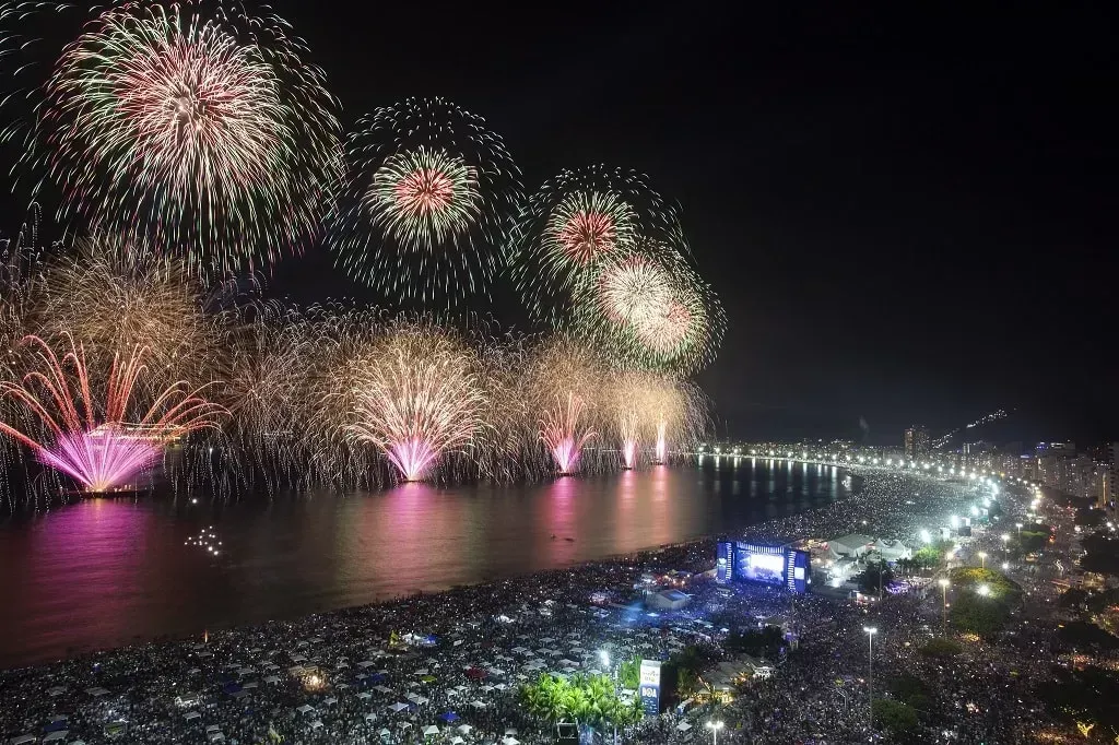 Fireworks exploding over a crowded beach at night; pink, white, and gold bursts. Rio de Janeiro