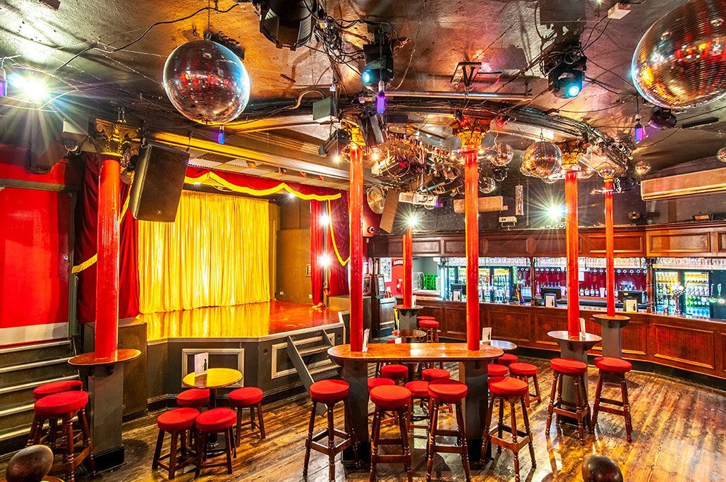Interior of a club with stage, bar, stools, red accents, and disco balls. Royal Vauxhall Tavern