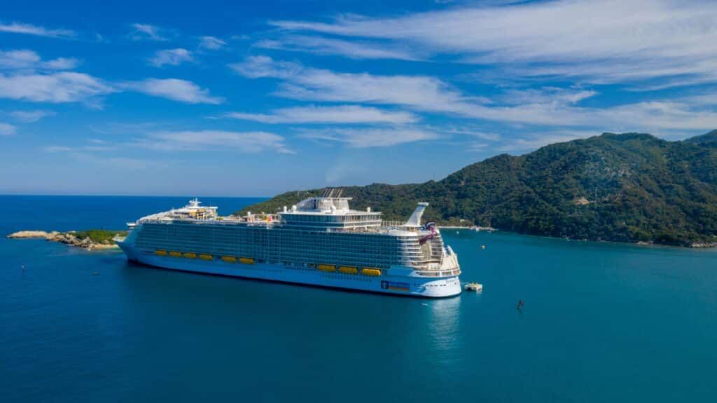 Large cruise ship docked in turquoise water near a forested island under a blue sky.