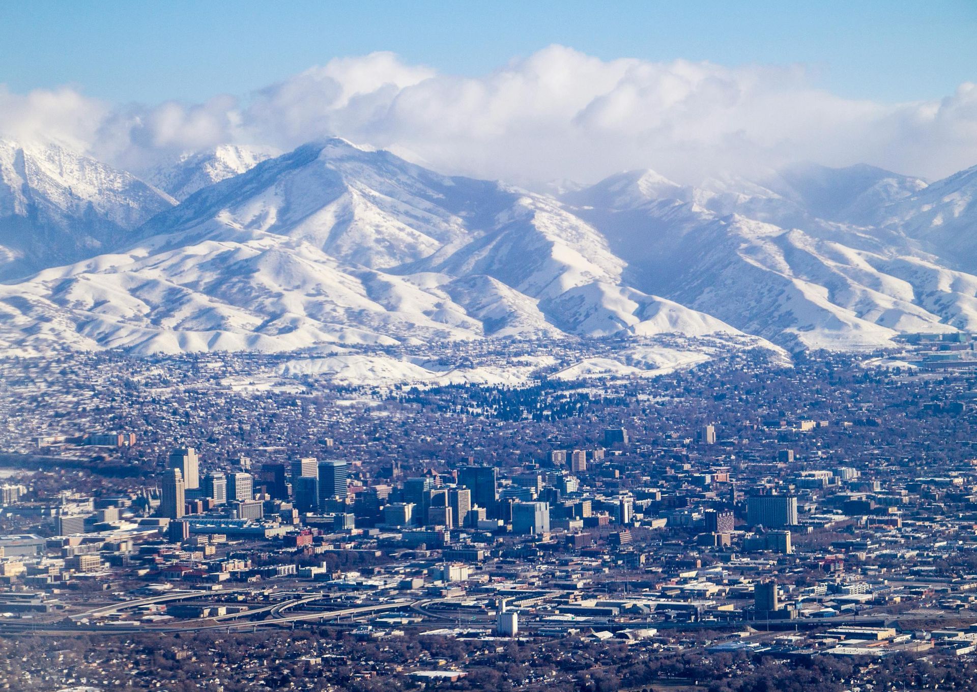 City skyline below snow-covered mountains, with white clouds in a bright blue sky.Salt Lake City, Utah
