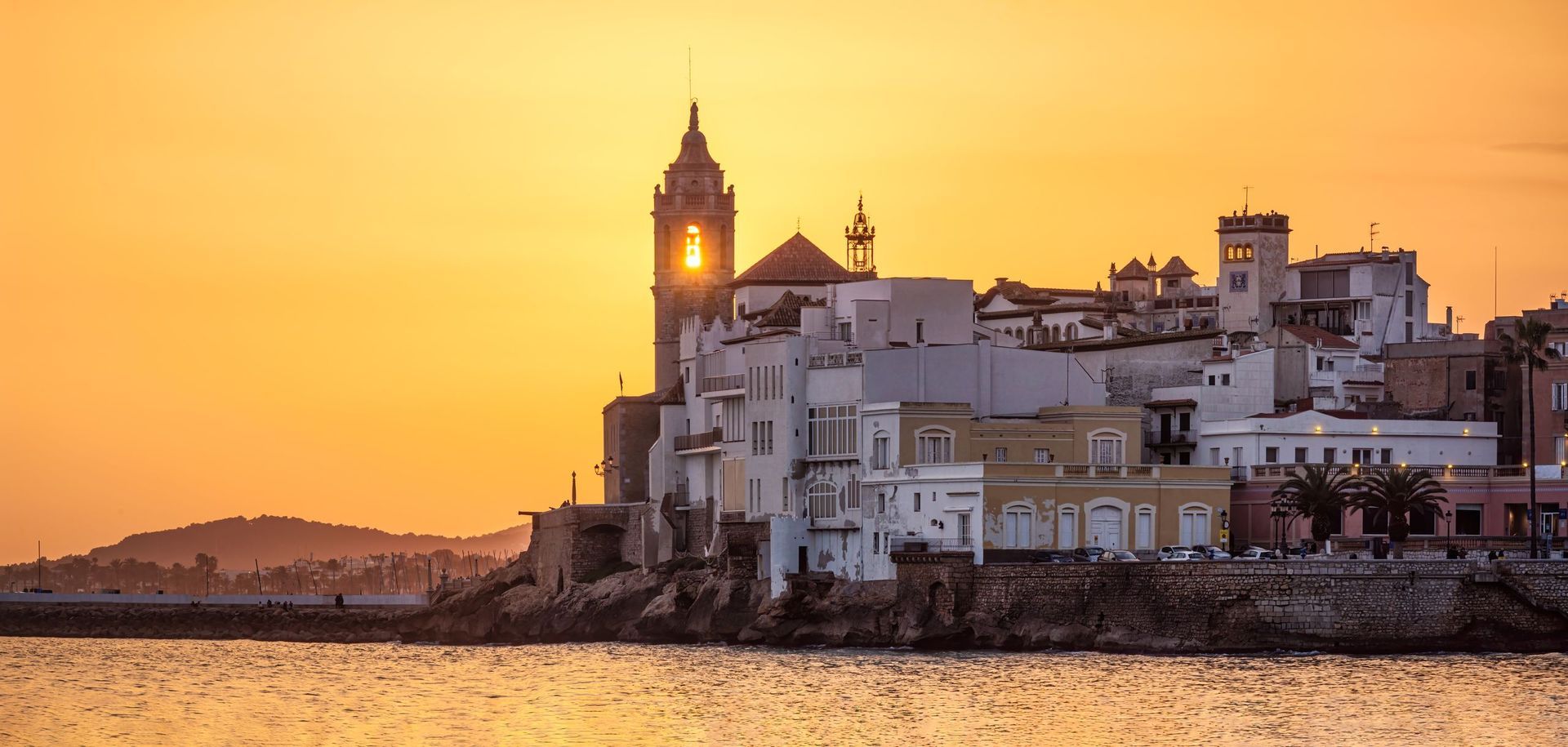 Sunset over a coastal city with a tall church tower, reflecting in the water.