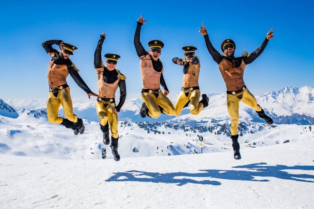 Five men in gold ski pants and military hats jump in the snow, arms raised, with a mountain backdrop.