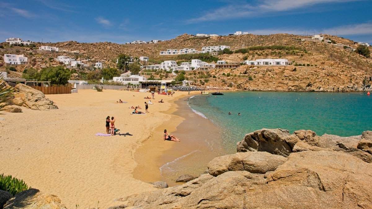 Beach scene with golden sand, turquoise water, and white buildings on a hillside, sunny day. Super Paradise Beach Mykonos