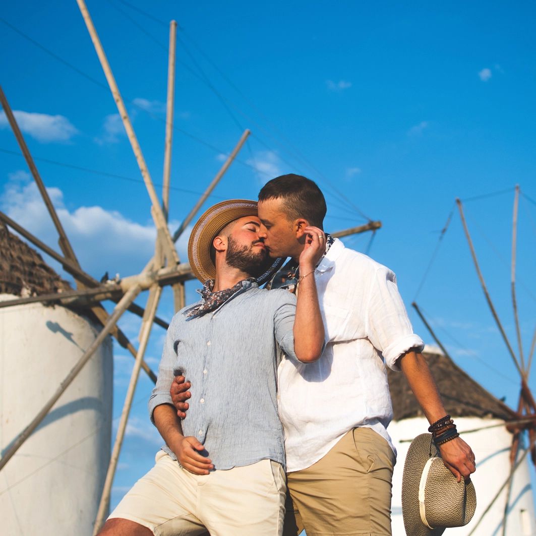 Two men kissing in front of white windmills and a blue sky. One wears a hat, the other holds one. Mykonos
