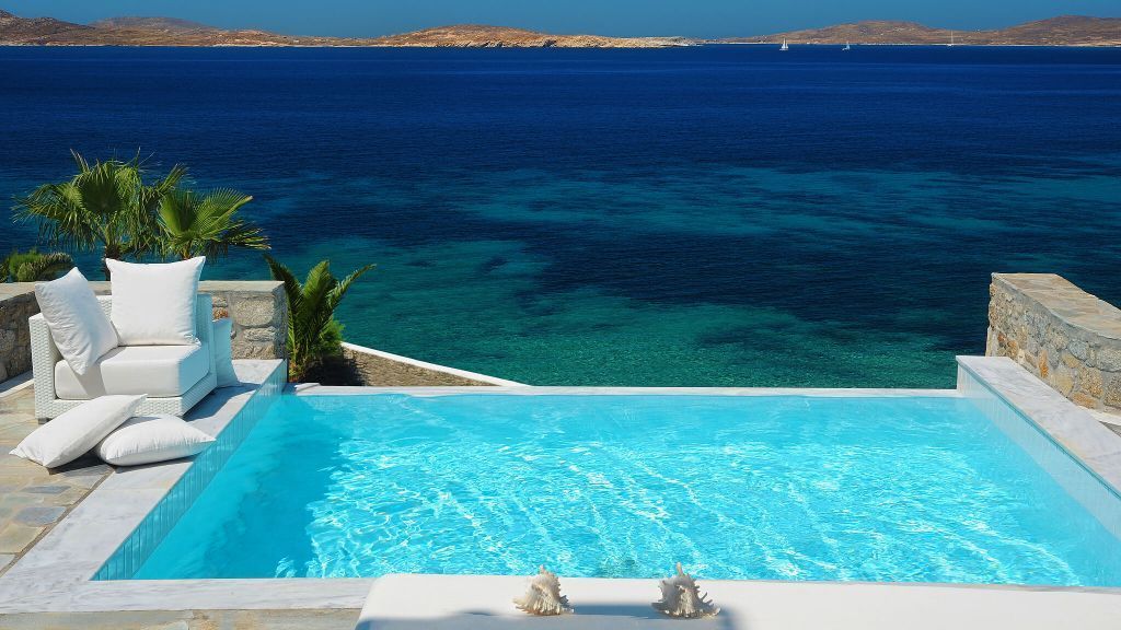 A blue swimming pool overlooking a deep blue sea and distant islands, with white lounge furniture.