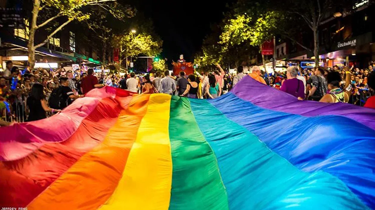 Large rainbow flag carried by a crowd in a city street at night during a Pride celebration. Sydney Mardi Gras