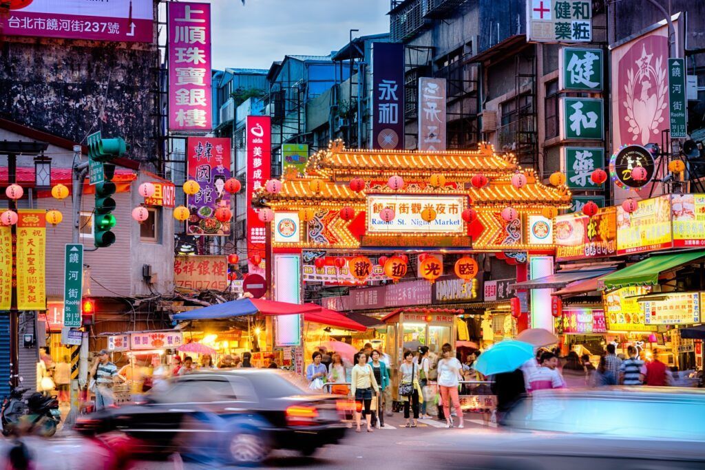 Busy Asian street scene with a decorative archway, colorful signs, and a blur of people and traffic at dusk. Taiwan