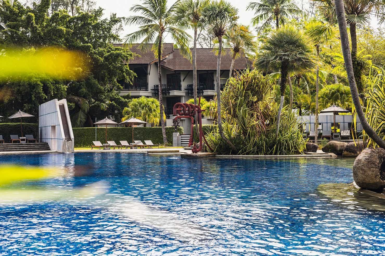 Swimming pool at a resort with a building and palm trees in the background. The The Slate - Phuket