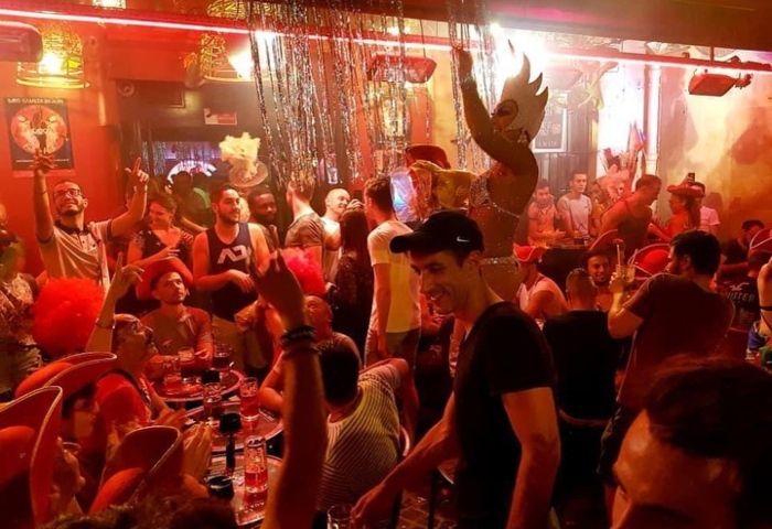 Crowded bar scene with people dancing and a performer on a platform. Red and orange lighting.