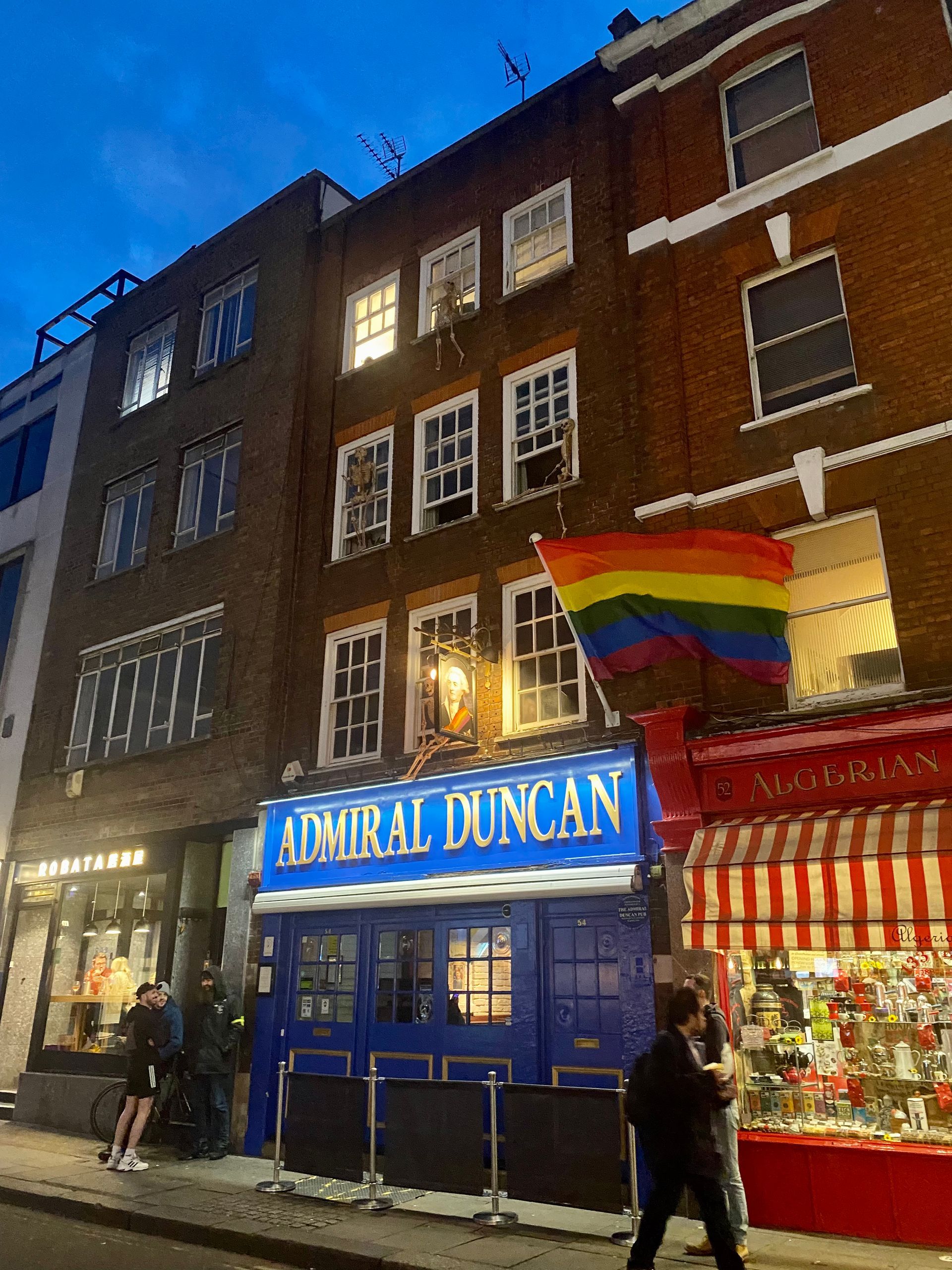 Admiral Duncan pub with rainbow flag, blue facade, brick buildings, and street scene at dusk.