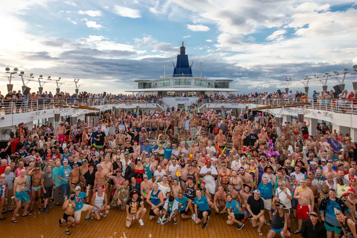Large group of people on cruise ship deck; many are in swimwear, posing for photo with ship structure in background. Vacay_Cruise